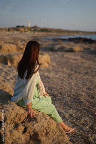 Pensive young woman sitting on a rocky shore and watching the sea during a beautiful golden sunset