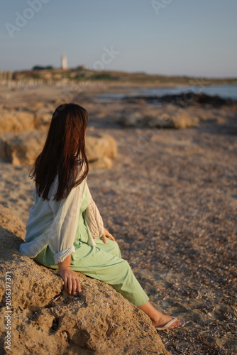 Pensive young woman sitting on a rocky shore and watching the sea during a beautiful golden sunset