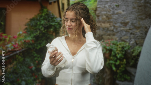 фотографія Young woman holding a plastic water bottle, touching temple with hand and grimacing near a stone building and garden; discomfort dehydration