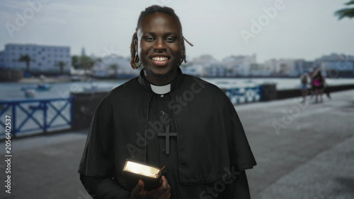 Obraz na plátně Man priest holding glowing bible on seaside promenade near harbor railing, smiling and displaying cross necklace; serenity faith