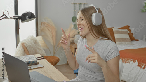 Bild auf Leinwand Young student woman tapping fingers on laptop while smiling, headphones on, books and mug at desk in bedroom study nook; focus joy