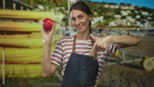 Quadro em tela Woman holds red apple with gloved hand and points finger to apple on beach; healthy eating cheerful