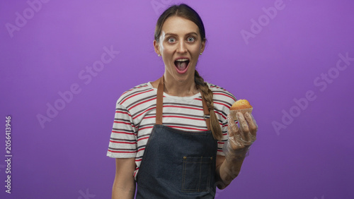 Tableau sur toile Woman holding muffin with gloved hand, open mouth, wearing apron and striped shirt in purple studio; surprise moment