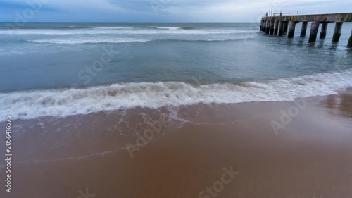 Waves gently lapping on a sandy beach with a distant pier under a cloudy sky.