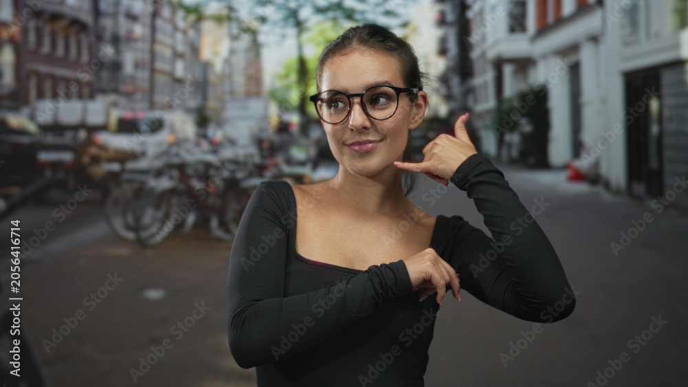 Fototapeta premium Woman with glasses making call me hand gesture on a city street lined with bicycles and brick buildings wearing a black long sleeve shirt; playful flirtation.