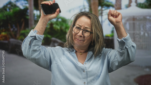 Fotografija Hispanic woman holding smartphone and raising arms while singing with head tilted on a street lined with palms and a fountain; joy celebration