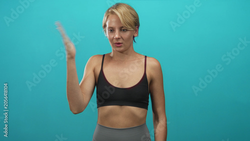 Fotografie Woman blond pinches nose with hand and grimaces while wearing sports bra and leggings in studio turquoise backdrop; disgust aversion