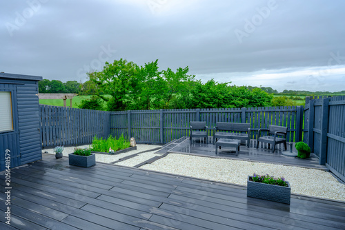 Modern deck with dark wood planks and gravel patio. Garden chairs sit invitingly near a low fence. Overcast sky adds a calm, tranquil mood. Greenery frames the outdoor space with natural beauty
