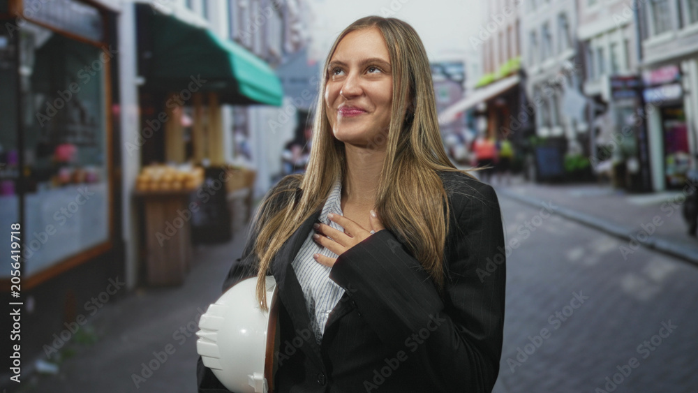 Fototapeta premium Young hispanic woman holding white hardhat waving hand on an urban street with storefronts; gratitude ambition.