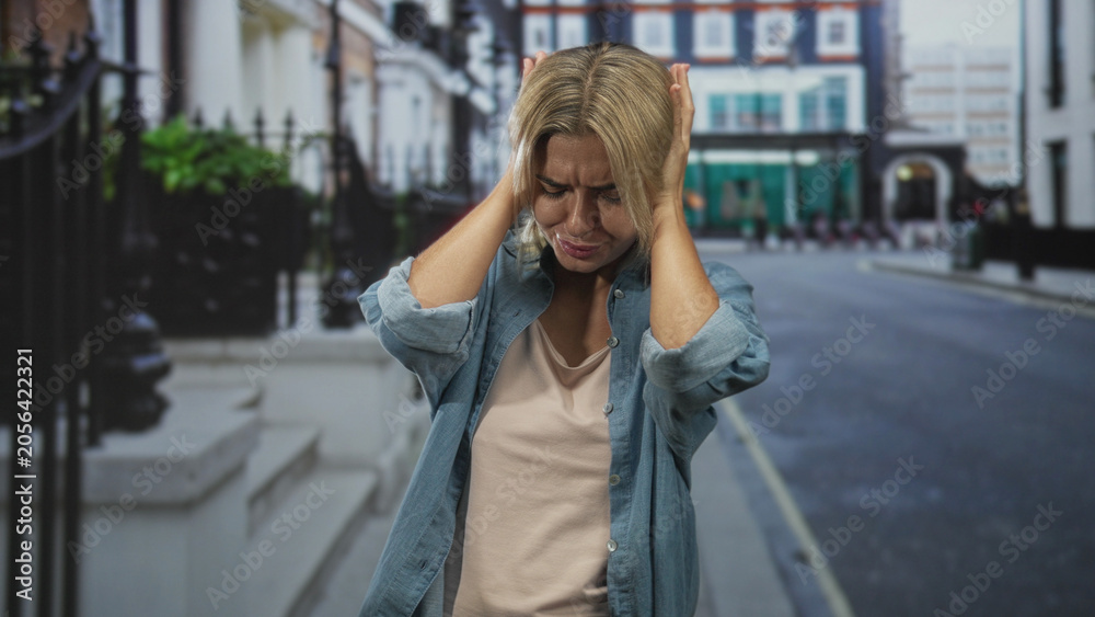 Fototapeta premium Woman covering ears with hands on a city street, denim shirt visible, wincing from headache; anxiety isolation.