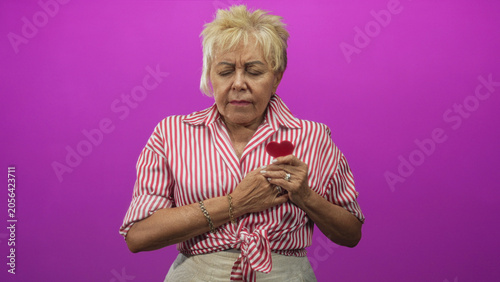 Bild auf Leinwand Senior woman holding a small red felt heart to chest with hands, wearing red striped shirt in studio; nostalgia memory love