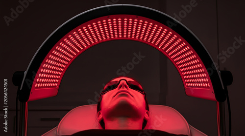 Low angle medium shot of a woman lying under an arched red led light therapy panel for facial skin rejuvenation treatment in a professional aesthetic clinic setting
