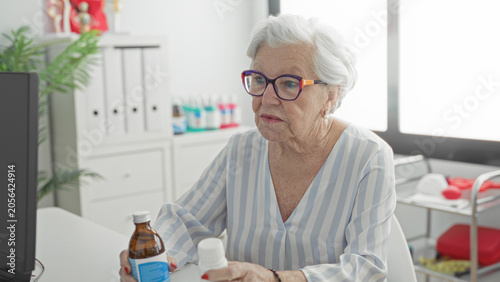 Εκτύπωση καμβά Elderly woman with glasses holds a medicine bottle and opens the cap with her hand while seated at a clinic desk, intently reading the label; concern medication safety and care