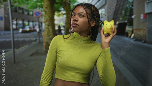 Photos Woman holding yellow piggybank on a busy city street near tree with raised eyebrow and hand extended; financial skepticism