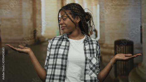 Obraz na plátně Woman shrugging palms up on old town street by stone columns, wearing white t shirt and plaid shirt with hands extended to sides; confident decision