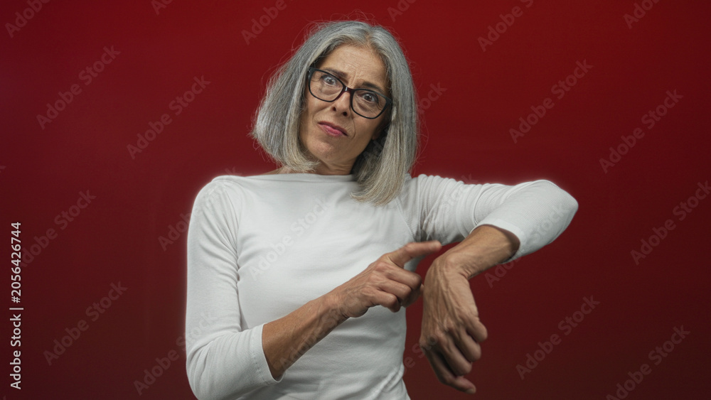 Fototapeta premium Middle age grey haired woman in white shirt pointing finger to wrist in studio with red wall; time impatience.