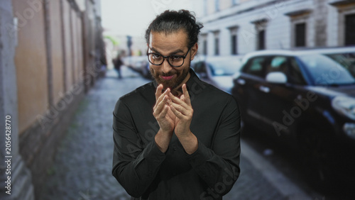 Φωτογραφία Man with glasses rubbing fingers together in black shirt on cobblestone street; scheming slyness cunning intent