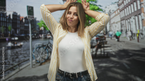 Obraz na plátně Woman making bunny ears with hands on a canal street, wearing white shirt and jeans with belt; playful