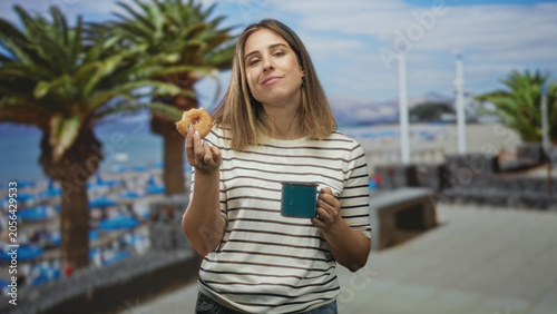 Foto Woman holding a glazed donut and blue mug, tongue out and smiling in a striped shirt on a street boardwalk by palm trees and beach umbrellas; playful relaxation