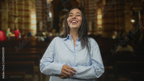 Foto Woman laughing with face toward camera, hands clasped over stomach inside a catholic church nave with wooden pews and stone arches; joy faith