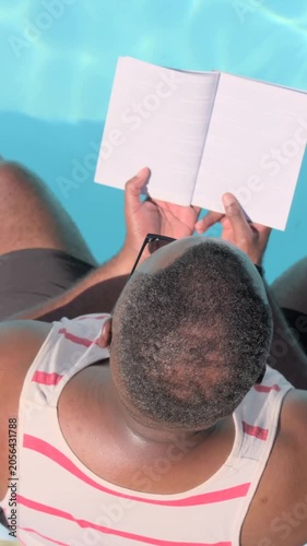 Vertical video: Adjusting man in striped tank holding open notebook to read at pool, sunglasses