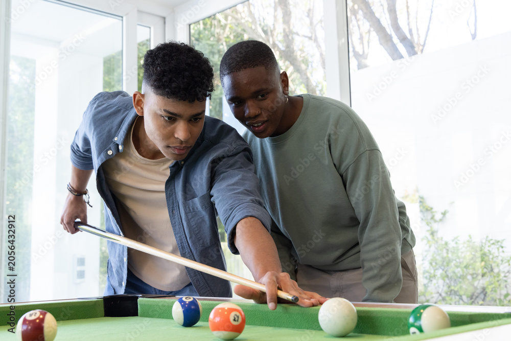Obraz premium Hispanic and African American teenage males lining up shot on pool table at home with cue