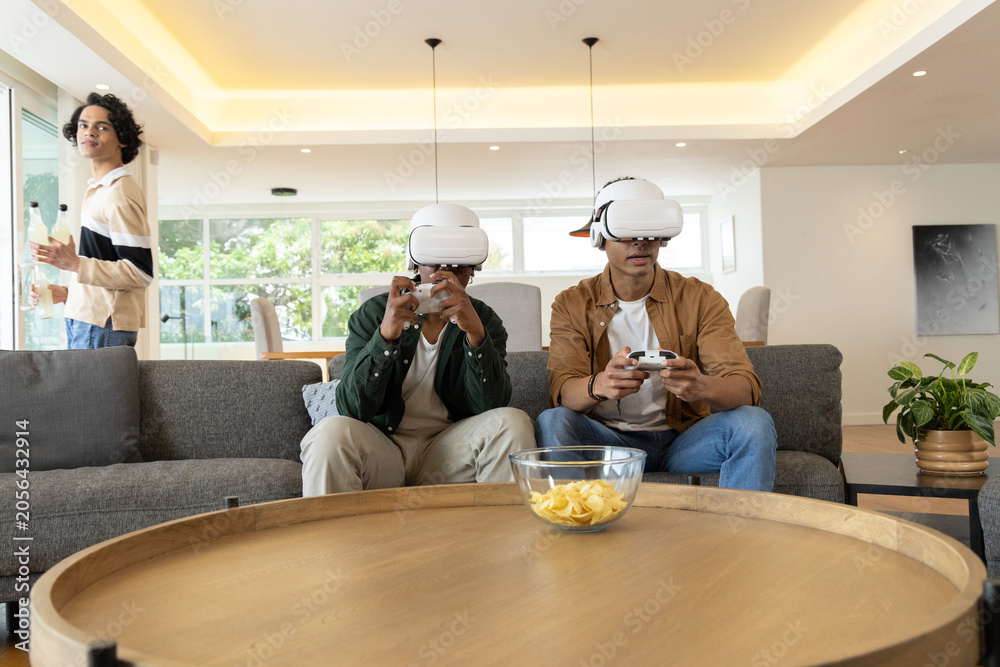 Obraz premium Diverse teenage males playing VR games on gray sofa wearing white headsets and holding controllers