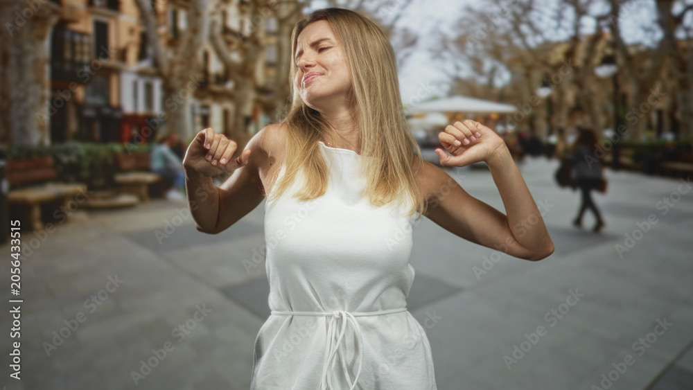 Fototapeta premium Blonde woman stretching and yawning with hands behind head showing bare arms on a street; sleepiness morning routine.