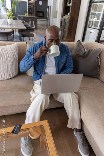 Senior African American man sitting on sofa at home, wearing denim, holding mug and using laptop