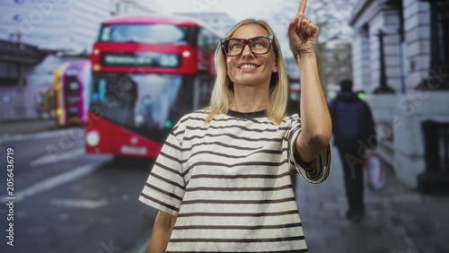 Photos Woman smiling and pointing index finger upward on a busy city street beside a red doubledecker bus and walking pedestrians; delight curiosity wonder
