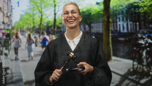 Obraz na plátně Woman judge in black robe holding gavel and soundblock, winks and raises gavel on street; confidence justice