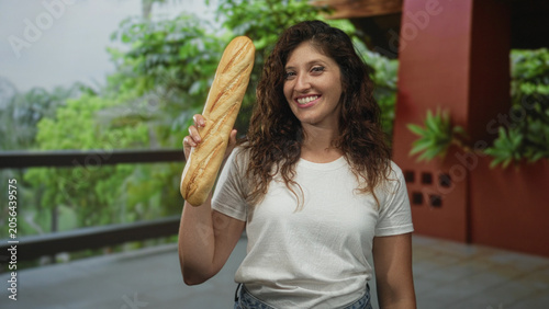 Obraz na plátně Young hispanic woman smiles and holds a baguette aloft in right hand on hotel balcony inside building; joy friendly warmth
