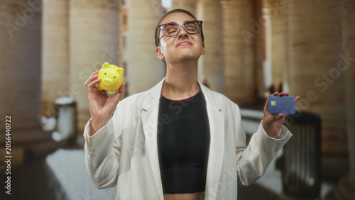 Tableau sur toile Woman with short hair holds yellow piggy bank in left hand and blue creditcard in right hand, hand slightly raised inside historic columned building; confidence saving money