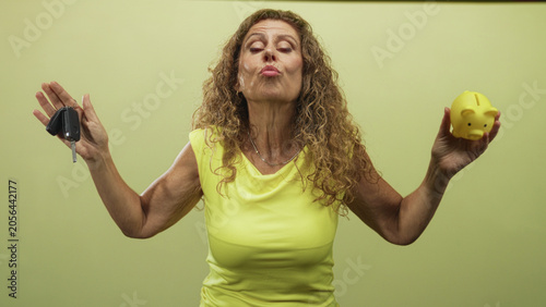 Obraz na plátne Middle aged hispanic woman holding car keys and a yellow piggy bank, bare hands raised and puckering lips in studio; financial choice playful