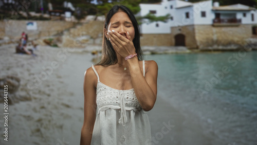 Cuadro en lienzo Thai woman covers mouth with hand by waterfront building in white sundress and bracelet, showing a yawn and closed eyes; fatigue