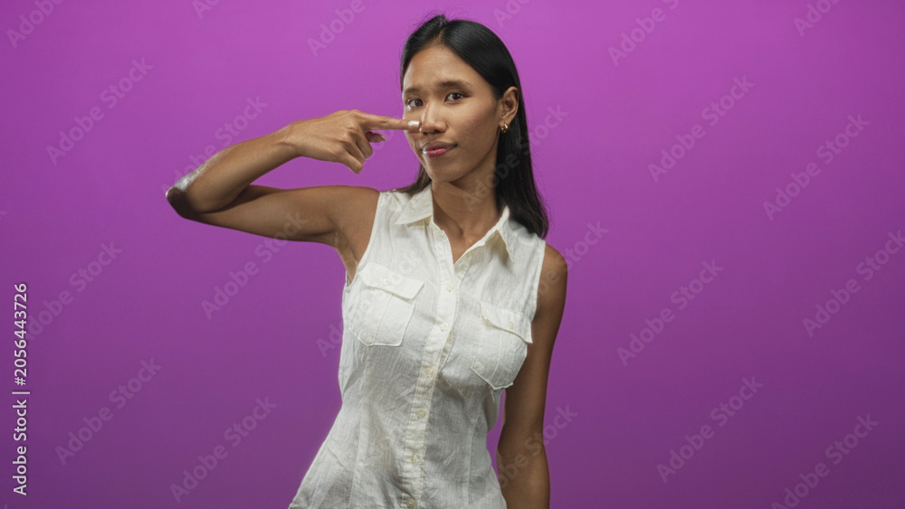 Obraz premium Thai woman points finger to nose in studio with purple backdrop and sleeveless white shirt; playful curiosity.