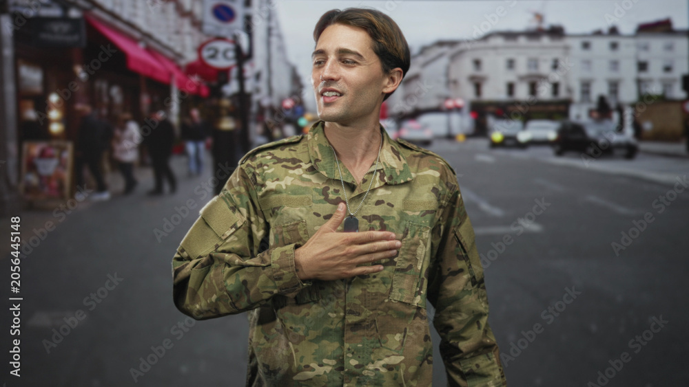 Fototapeta premium Man soldier in camouflage with hand on chest and dog tag visible on street, touching mouth mid gesture; pride service duty.