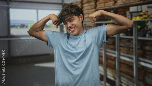 Teenage boy flexes biceps and sticks out tongue in a greenhouse building, beside metal shelves and wooden crates with potted flowers; playful confidence.
