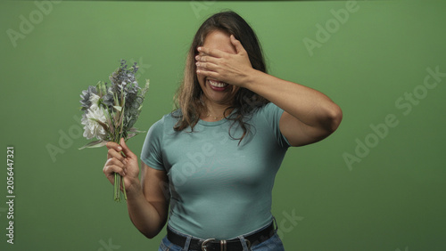 Obraz na plátně Woman holding bouquet of white and muted blooms, covering her eyes with one hand in green studio; shyness