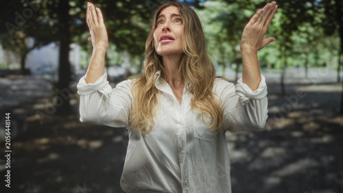 Fotografie Woman with hands raised in pleading gesture wearing white shirt on a tree lined street, teary expression and visible tears; pleading distress