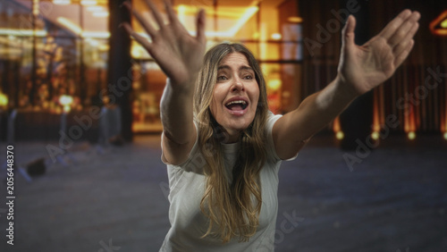 Foto Woman raising hands with open mouth reaching forward, visible hands and forearms, standing in a building near stanchions and warm lights; pleading hope
