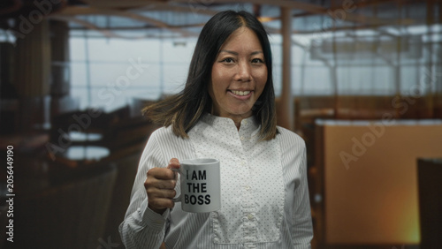 Quadro su tela Woman holds mug that reads i am the boss in restaurant building while smiling and extending the mug forward; quiet confidence