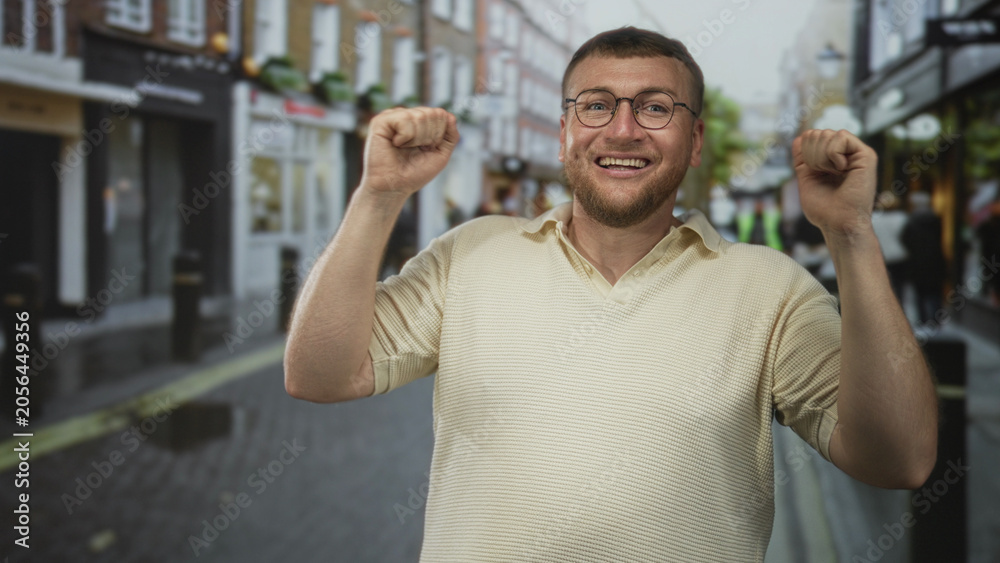 Naklejka premium Man smiling and fist pumping amid pedestrians on a busy urban street with shops and bollards; simple joy.