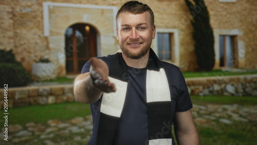 Fotografi Man young and stocky wearing black and white scarf offers handshake and presses hand to mouth at building entrance outdoors; friendship welcome support