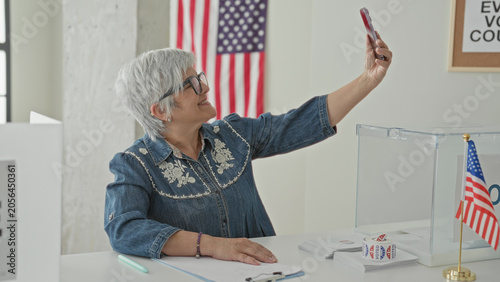 Konstfotografi Woman takes selfie with smartphone and raised arm at polling building beside clear ballot box and american flag; pride duty