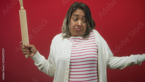 Canvastavla Hispanic woman holds a rolling pin aloft with one hand and shrugs shoulders, unimpressed face, striped shirt and cardigan in a studio with red backdrop; resignation kitchen humor