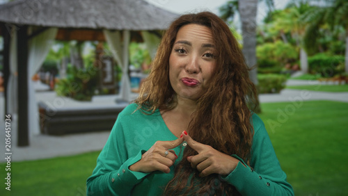 Fényképezés Woman touching index fingers together at a resort cabana near pool wearing green sweater and red nails; hesitant reflection decision