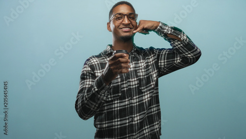 Tableau sur toile Young black man wearing glasses and plaid shirt holding takeaway coffee cup making call me gesture and smiling in studio; cheerful connection