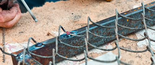 Tableau sur toile Worker's dirty hand holding a welding electrode, joining metal rods to reinforce a rust-colored steel frame, capturing the skill and craft of metalworking