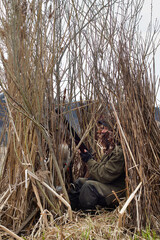 A duck hunter sits with a shotgun in his hands in a reed duck blind